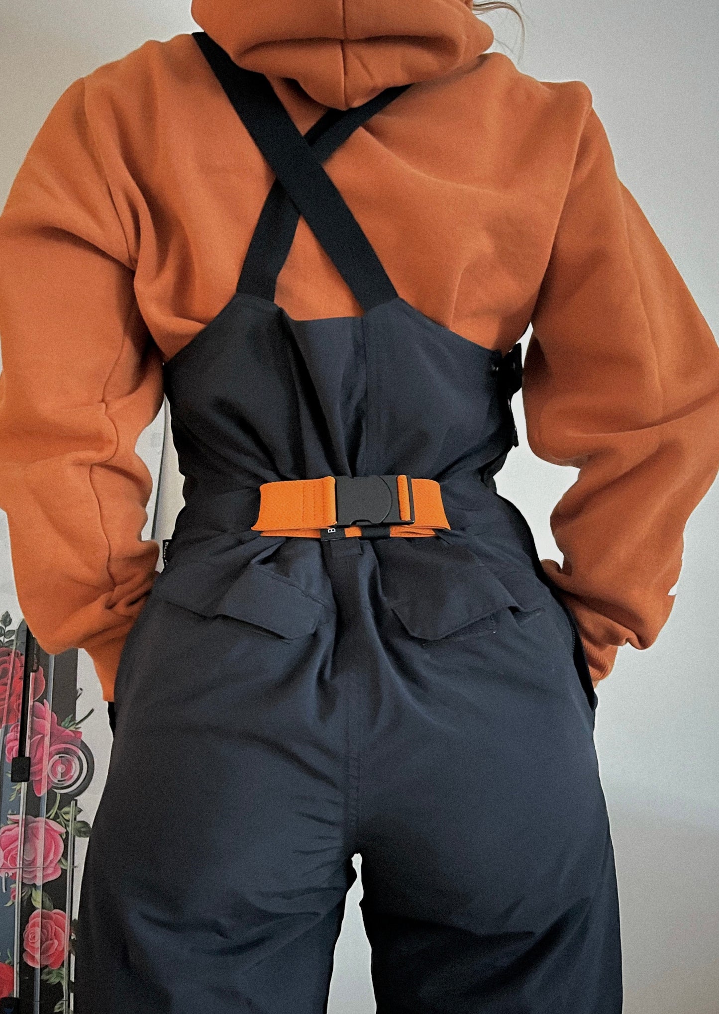 a girl and her snowboard facing backward wearing a black ski bib and a burnt orange bib belt